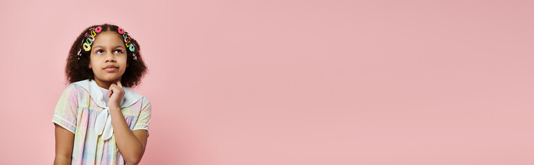 A cute African American girl with colorful hair clips stands against a pink background, thoughtfully gazing upwards.