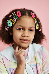 A young African American girl with colorful hair clips stands against a pink background.