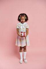 A young African American girl in a colorful dress holds a small gift box while standing against a pink background.