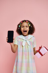 A young African American girl in a colorful dress smiles and holds a phone with a blank screen.