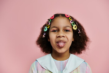 A Black girl with colorful hair clips sticks out her tongue and smiles playfully in front of a pink background.