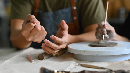 Close-up of hands working with clay on a pottery wheel. One person shapes a piece of clay while the other adds details with a sculpting tool. Both are focused on the intricate crafting process