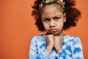 A young African American girl with curly hair stands against an orange background, wearing a trendy...