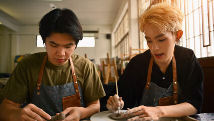 Two art students working on pottery projects in a studio. Both are focused, wearing aprons, and shaping clay with their hands and tools. The workspace is filled with natural light and art supplies