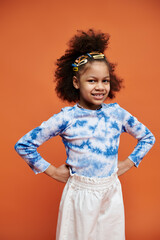 A young African American girl with colorful hair clips stands confidently against an orange background, wearing a trendy tie-dye shirt.