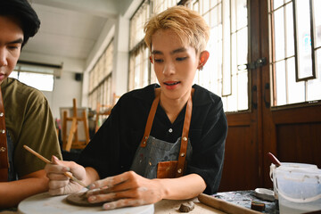 Two art students, one with blonde hair, work together on a pottery project. They are focused and...
