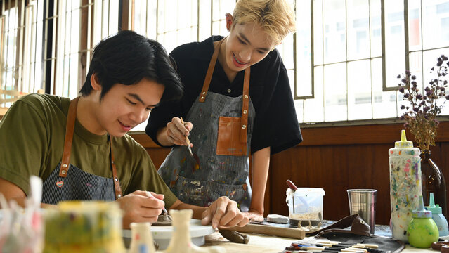 An art teacher assists a student in sculpting clay at a pottery wheel. Both are focused and smiling in a bright studio filled with art tools, paint supplies, and natural light from large windows