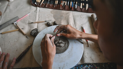 An overhead view of hands shaping a clay piece on a pottery wheel, surrounded by various sculpting tools and clay pieces, Crafts and DIY with people concept