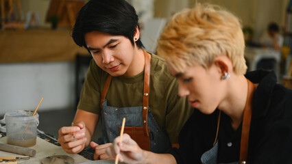 Close-up of An art instructor guides an LGBT student as they paint a ceramic piece. Both are...