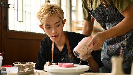 An instructor uses a hair dryer to help an LGBT student dry a painted ceramic piece. Both are focused and engaged in the creative process in a well-equipped, bright studio, Crafts and DIY concept