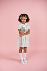 A young African American girl with colorful hair clips stands in a cute dress on a pink background.