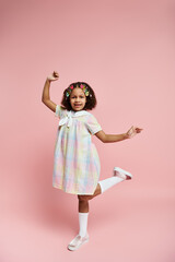 A joyful African American girl in a colorful dress and hair clips smiles for the camera against a pink background.
