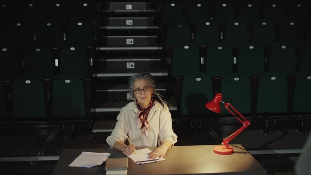 Experienced female casting director watching actor performing on stage and writing down notes during audition in theatre