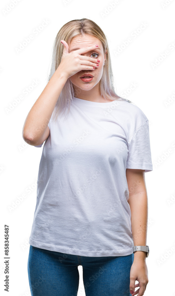 Wall mural Young blonde woman over isolated background peeking in shock covering face and eyes with hand, looking through fingers with embarrassed expression.