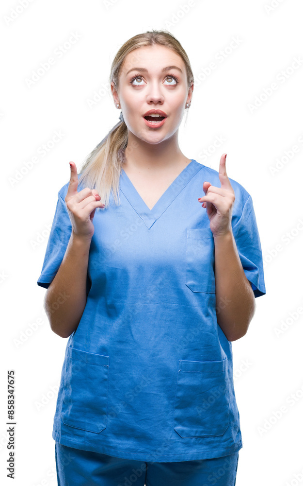 Sticker Young blonde surgeon doctor woman wearing medical uniform over isolated background amazed and surprised looking up and pointing with fingers and raised arms.