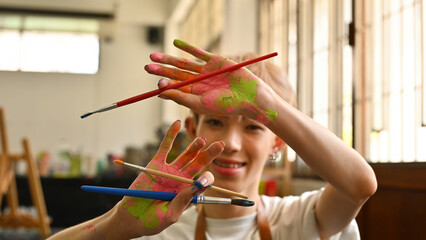 A joyful LGBT teenage artist displays their paint-covered hands and brushes, radiating creativity and happiness. The vibrant colors on their hands symbolize the passion and playfulness in their art