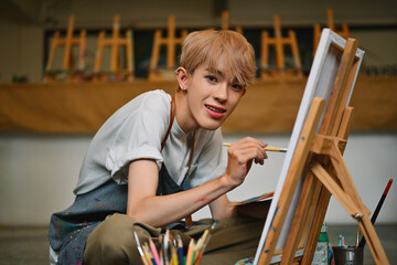 A joyful LGBT artist with blonde hair, wearing a paint-splattered striped apron, paints on a canvas in a bright, spacious art studio filled with light, easels, and various art supplies