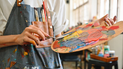 Close-up of an LGBT artist's hands mixing vibrant paints on a palette while wearing a striped apron...
