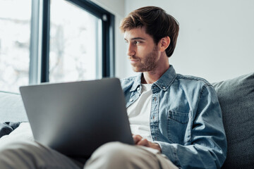 Handsome serious man sits on sofa with computer on laps, spend time at home web surfing information, use modern tech, make order on internet. Freelancing, telework, new software learning, tech concept