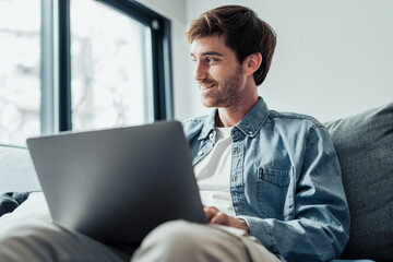 Handsome serious man sits on sofa with computer on laps, spend time at home web surfing information, use modern tech, make order on internet. Freelancing, telework, new software learning, tech concept