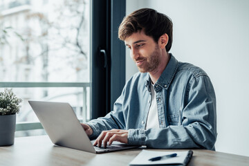 Side view handsome young businessman working with computer remotely, sitting at wooden table in office. Pleasant happy man communicating in social network, searching information online..