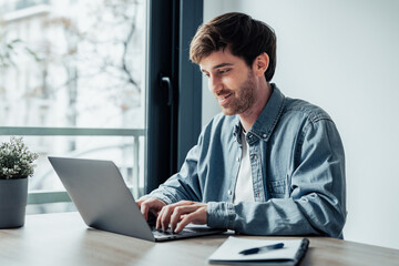Side view handsome young businessman working with computer remotely, sitting at wooden table in office. Pleasant happy man communicating in social network, searching information online..
