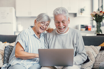 Pretty elderly 70s grey-haired couple resting on couch in living room hold on lap laptop watching movie smiling enjoy free time, older generation and modern wireless technology advanced users concept.