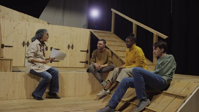 Senior female drama teacher holding papers and explaining script to young actors sitting on theatre stage