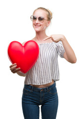 Blonde teenager woman holding red heart very happy pointing with hand and finger