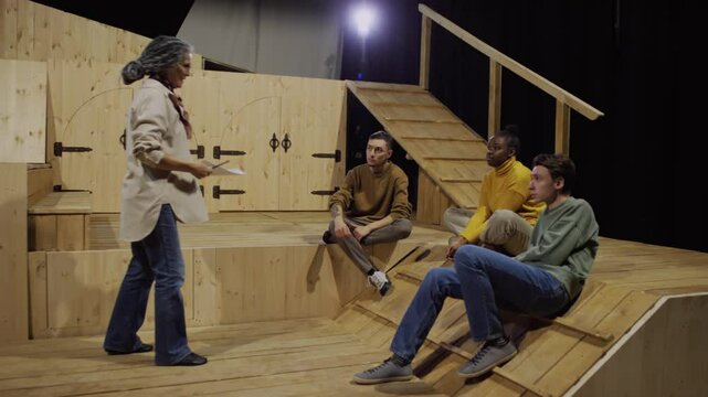 Mature female drama teacher holding script and explaining acting technique to you g diverse students sitting on theatre stage and listening attentively