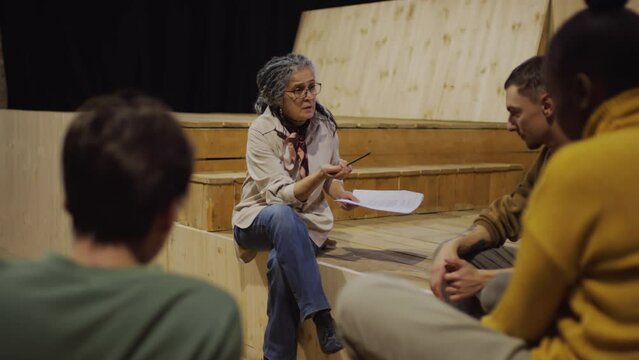 Mature female director sitting on stage in theatre and discussing script with group of young actors during rehearsal of play