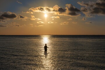 Fototapeta premium Person standing in the ocean water during a beautiful sunset with clouds in the sky