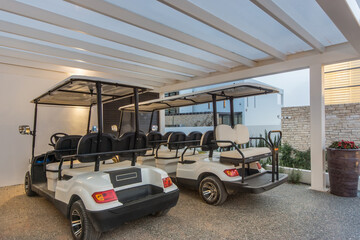 Two white golf carts parked under a white pergola at a resort. The golf carts are parked on a gravel driveway, with a stone wall and some greenery visible in the background. 