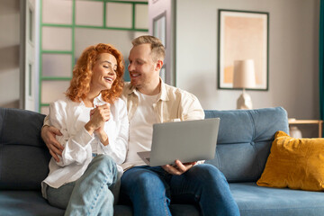 Young spouses shopping online on laptop, sitting on couch in living room interior. Sale for shopaholic, internet offer, video call and modern gadget