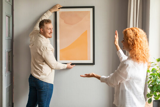 Young spouses arranging new flat, hanging up painting on wall. New home and relocation concept