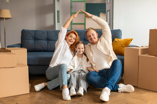 Happy young parents and kid promoting mortgage, real estate property buying, sitting on floor in new apartment with boxes behind, making hand roof gesture