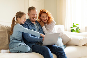 Cheerful family of three using laptop computer, daughter pointing at screen,shopping online while sitting on couch together. Technology and domestic lifestyle concept