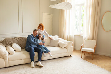 Obraz premium Loving parents and their daughter having video call on smartphone, resting and posing sitting on couch at home interior with free space