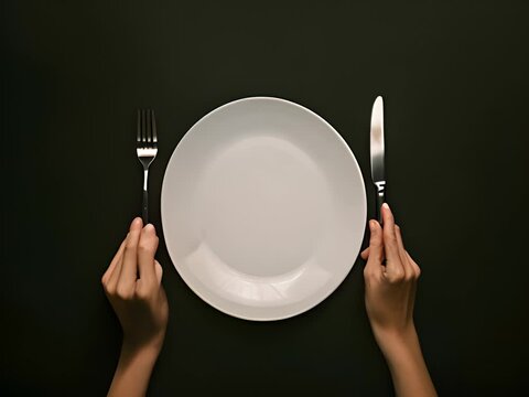 hands holding a knife and fork on a black background, an empty white plate.View from above.