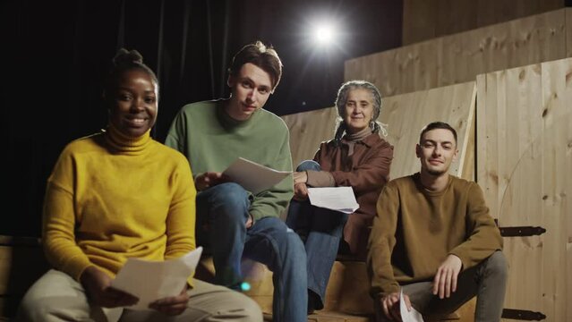 Group portrait of diverse actors and actresses sitting on stage, holding scripts and posing together for camera with smile during theatre rehearsal