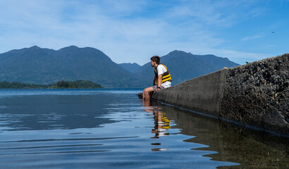 Picture-perfect scene of a young man seated on the lake dock, taking in the stunning vista during a delightful summer afternoon.