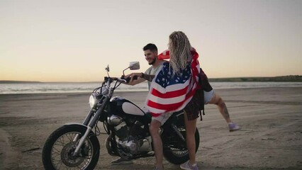 Woman in love running to motorcyclist guy along the beach at sunset, wrapped in an American flag - Powered by Adobe