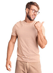 Young caucasian man wearing casual clothes and glasses smiling with happy face looking and pointing to the side with thumb up.