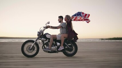 A couple in love with an American flag rides a motorcycle on the beach during sunset, enjoying a moment of freedom - Powered by Adobe