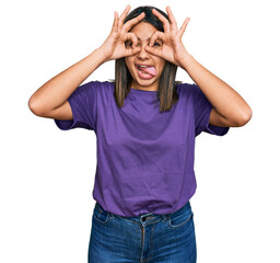 Young hispanic girl wearing casual purple t shirt doing ok gesture like binoculars sticking tongue out, eyes looking through fingers. crazy expression.