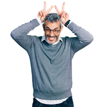 Middle age hispanic with grey hair wearing glasses posing funny and crazy with fingers on head as bunny ears, smiling cheerful