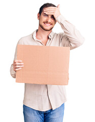 Young handsome caucasian man holding empty cardboard banner stressed and frustrated with hand on head, surprised and angry face