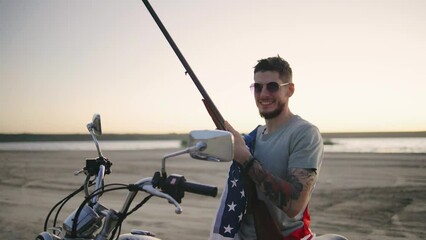 A man in sunglasses wripped in american flag sitting on motorcycle and rifle at sunset beach, embodying adventure, freedom, and a rugged, macho attitude in a scenic landscape - Powered by Adobe