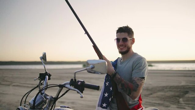 A man in sunglasses wripped in american flag sitting on motorcycle and rifle at sunset beach, embodying adventure, freedom, and a rugged, macho attitude in a scenic landscape
