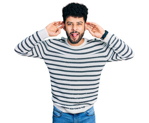 Naklejka premium Young arab man with beard wearing casual striped sweater smiling pulling ears with fingers, funny gesture. audition problem